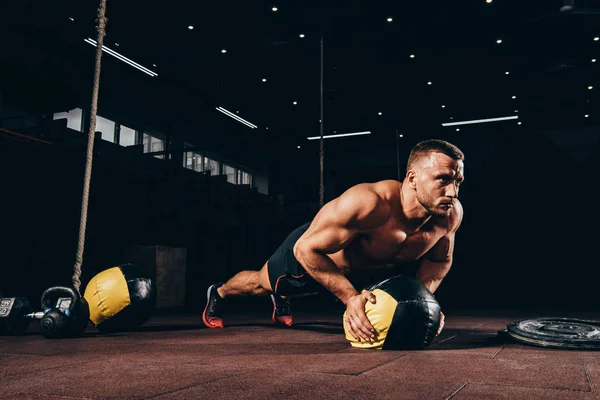 Apuesto deportista atlético haciendo flexiones en la pelota de la medicina en el gimnasio oscuro - foto de stock