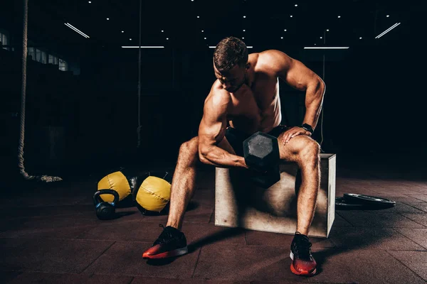 Schöner muskulöser Sportler, der die Hantel hebt, während er in der dunklen Turnhalle auf einem Würfel sitzt — Stockfoto