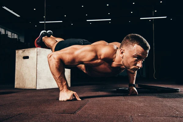 Handsome athletic sportsman doing push ups with feet on cube in dark gym — Stock Photo