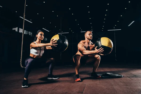 Deportista atlético y deportista haciendo ejercicio con bolas de medicina juntos en el gimnasio oscuro - foto de stock