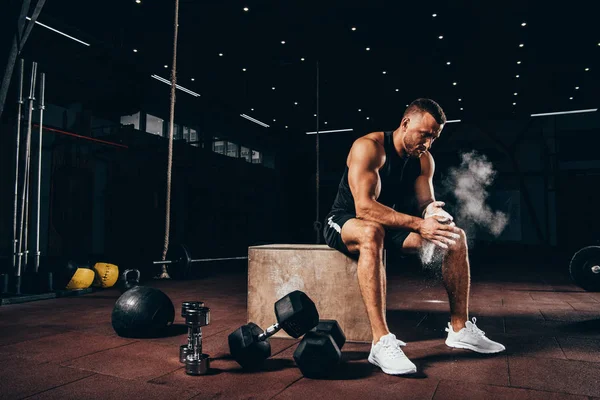 Handsome athletic man sitting on cube with gym equipment around and clapping hands with talc before workout — Stock Photo