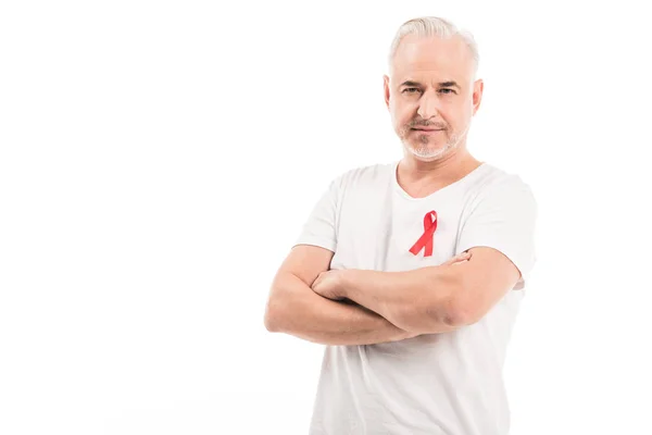 Красивый зрелый мужчина в пустой белой футболке со СПИДом осознание красной ленты глядя на камеру со скрещенными руками изолированы на белом — стоковое фото