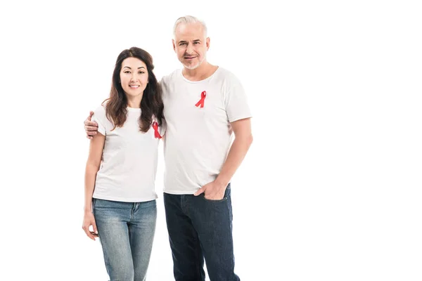 Smiling adult interracial couple in blank t-shirts with aids awareness red ribbons embracing and looking at camera isolated on white — Stock Photo