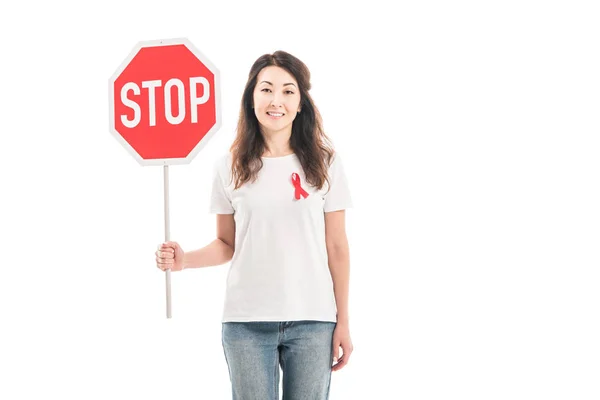 Smiling adult asian woman with aids awareness red ribbon on t-shirt holding stop road sign isolated on white — Stock Photo