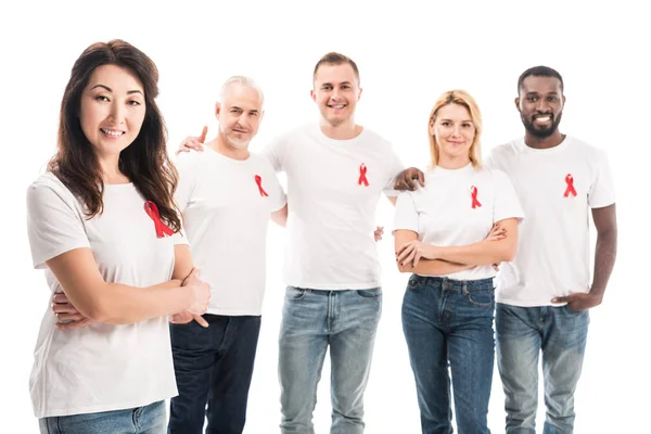 Sonriente mujer asiática con brazos cruzados mirando a la cámara con grupo de personas en blanco camisetas blancas con sida conciencia cintas rojas de pie aislado en blanco - foto de stock