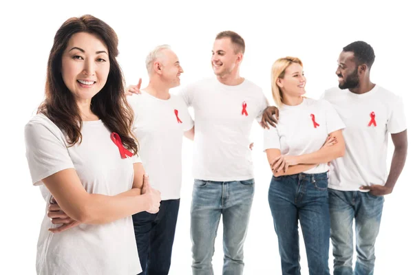 Happy asian woman with crossed arms looking at camera with group of people in blank white t-shirts with aids awareness red ribbons standing isolated on white — Stock Photo