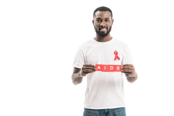 Glücklich afrikanisch-amerikanischer Mann in weißem T-Shirt mit Aids-Bewusstsein rotes Band und Blöcke mit Aids-Schriftzug isoliert auf weiß — Stockfoto