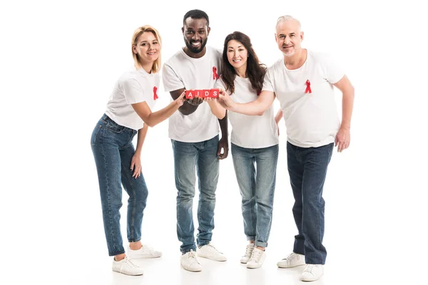 Smiling multiethnic people in blank white t-shirts with aids awareness red ribbons holding blocks with AIDS lettering isolated on white — Stock Photo