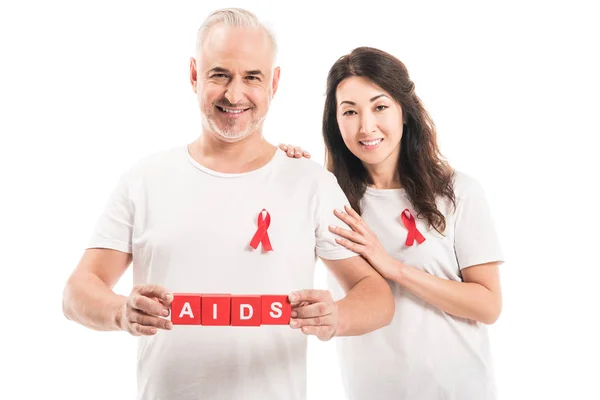 Smiling adult interracial couple in blank t-shirts with aids awareness red ribbons and blocks with AIDS lettering looking at camera isolated on white — Stock Photo