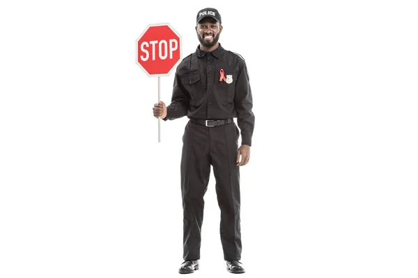 Happy african american police officer with aids awareness red ribbon and stop road sign isolated on white — Stock Photo