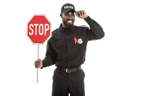 Sorridente policial afro-americano com aids conscientização fita vermelha e parar sinal de estrada isolado no branco — Fotografia de Stock