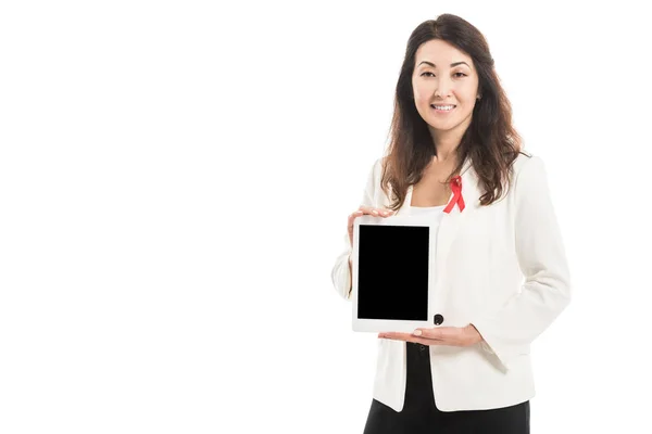 Smiling asian businesswoman with aids awareness red ribbon on jacket holding tablet and looking at camera isolated on white — Stock Photo