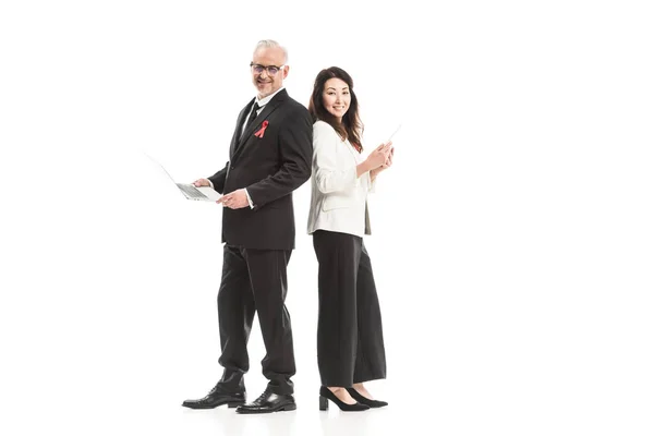 Successful adult businesspeople with aids awareness red ribbons working with gadgets while standing back to back and looking at camera isolated on white — Stock Photo