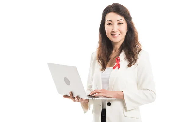 Happy asian businesswoman with aids awareness red ribbon on jacket using laptop and looking at camera isolated on white — Stock Photo
