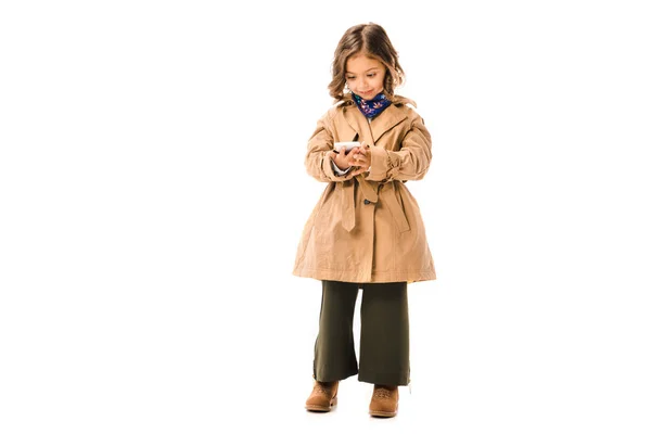 Hermoso niño pequeño en gabardina usando teléfono inteligente aislado en blanco — Stock Photo