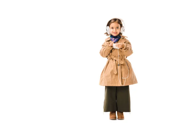 Adorable petit enfant dans un élégant trench coat écouter de la musique avec des écouteurs et en utilisant smartphone isolé sur blanc — Photo de stock