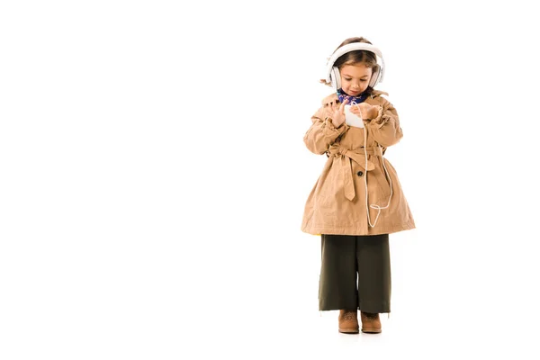 Hermoso niño pequeño en gabardina elegante escuchar música con auriculares y el uso de teléfono inteligente aislado en blanco - foto de stock