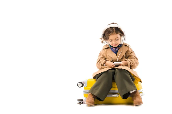 Adorable niño pequeño en gabardina escuchar música con auriculares y el uso de teléfono inteligente mientras está sentado en la maleta amarilla aislado en blanco — Stock Photo