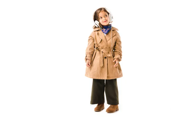 Beautiful little child in trench coat listening music with headphones isolated on white — Stock Photo