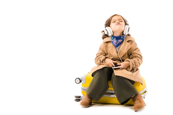 Criança sorrindo em casaco de trincheira ouvir música com fones de ouvido e sentado em mala amarela isolada em branco — Fotografia de Stock