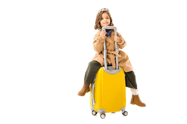 Beau petit enfant en trench coat assis sur valise jaune isolé sur blanc — Photo de stock