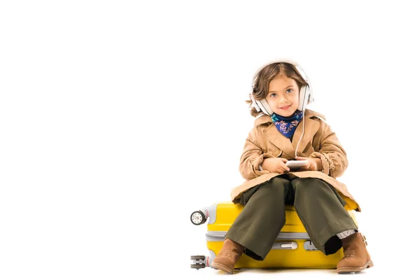 Hermoso niño pequeño en gabardina escuchando música con auriculares y sentado en la maleta amarilla aislado en blanco - foto de stock
