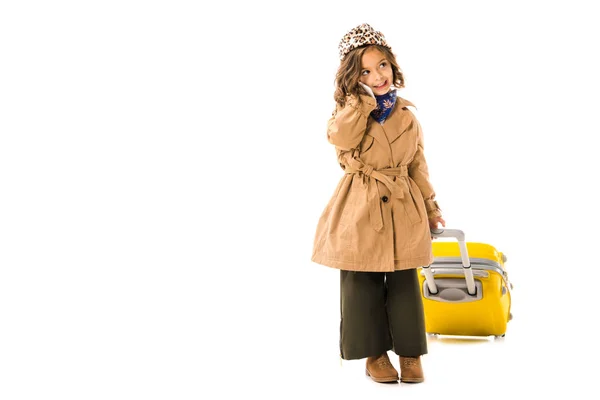 Happy little child in trench coat with yellow suitcase talking by phone isolated on white — Stock Photo