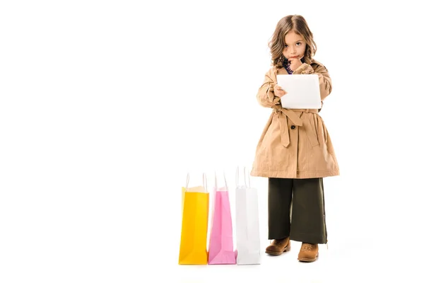 Hermoso niño pequeño en gabardina con coloridas bolsas de compras utilizando tableta aislada en blanco - foto de stock