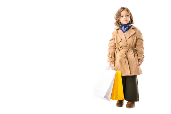 Adorable stylish child in trench coat with colorful shopping bags looking at camera isolated on white — Stock Photo