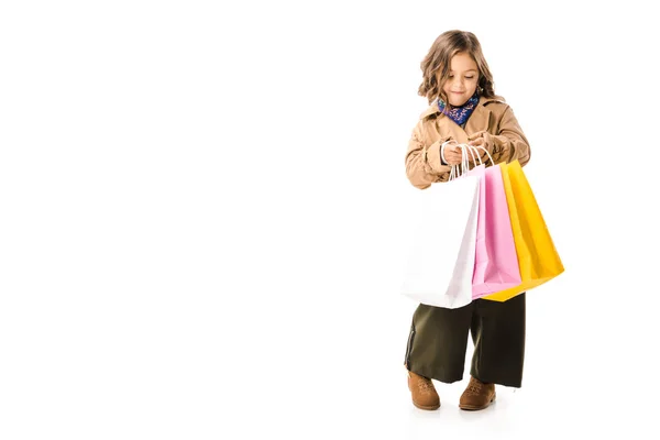 Schönes kleines Kind im Trenchcoat mit bunten Einkaufstaschen auf weißem Hintergrund — Stockfoto