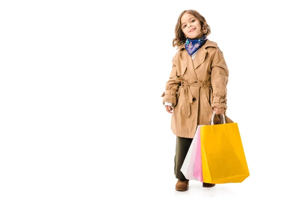 Entzückendes kleines Kind im Trenchcoat mit bunten Einkaufstaschen, das isoliert auf weiß in die Kamera blickt — Stockfoto