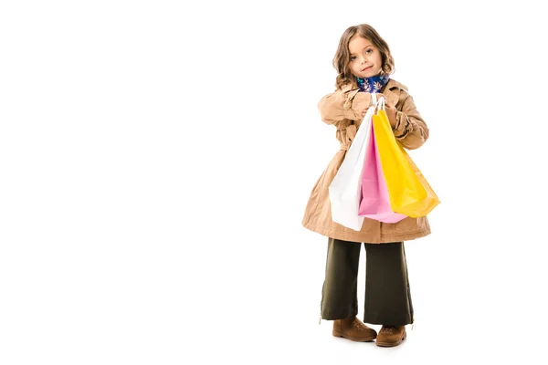 Niño pequeño con estilo en gabardina con coloridas bolsas de compras mirando a la cámara aislada en blanco - foto de stock