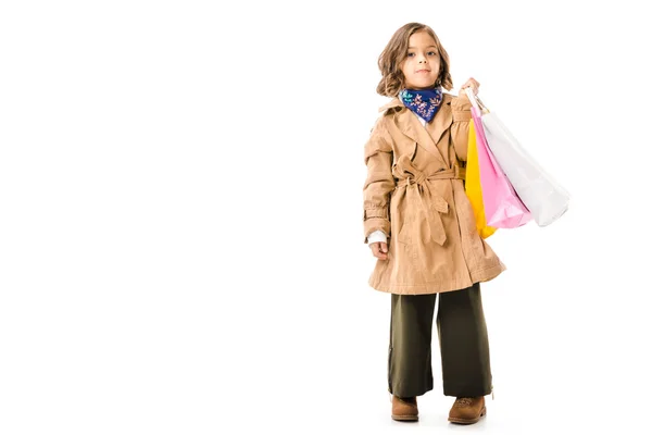 Niño pequeño con estilo en gabardina con coloridas bolsas de compras aisladas en blanco - foto de stock