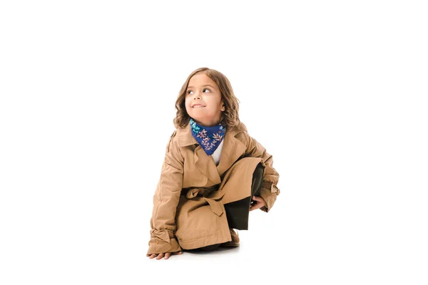Stylisches kleines Kind im Trenchcoat auf dem Boden sitzend isoliert auf weiß — Stockfoto