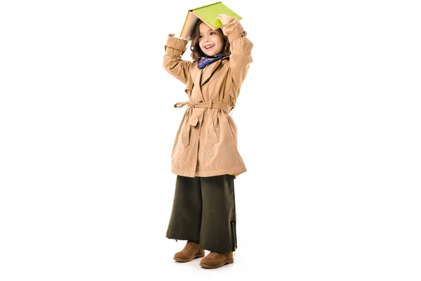 Beautiful little child in trench coat covering head with book isolated on white — Stock Photo