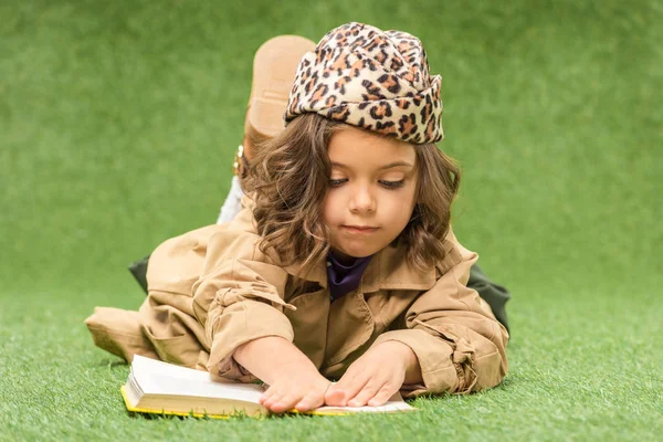 Stylish kid reading book while lying on green grass — Stock Photo