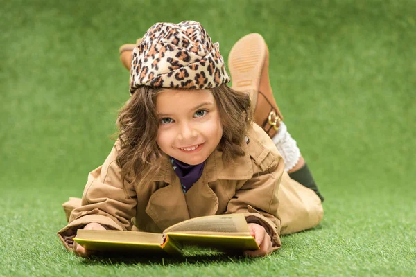 Stylish smiling kid with book looking at camera while lying on green grass — Stock Photo