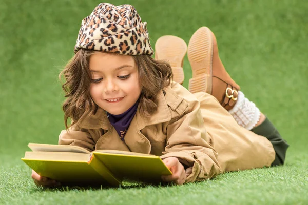 Stylish kid reading book while lying on green grass — Stock Photo