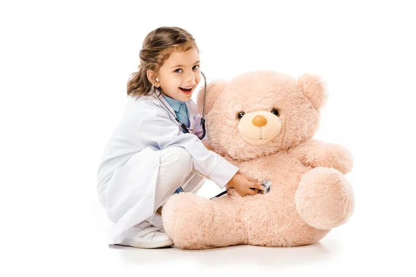 Niño vestido de médicos blanco abrigo con estetoscopio jugando con oso de peluche aislado en blanco - foto de stock