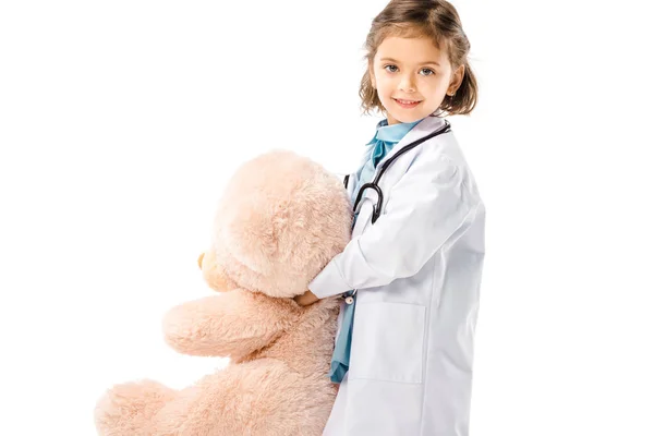 Lächelndes Kind im weißen Mantel mit Stethoskop, das einen großen Teddybär auf weißem Grund hält — Stockfoto