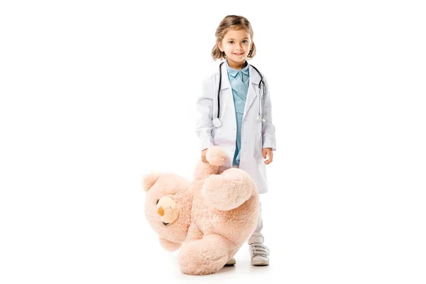 Kid dressed in doctors white coat with stethoscope holding big teddy bear isolated on white — Stock Photo