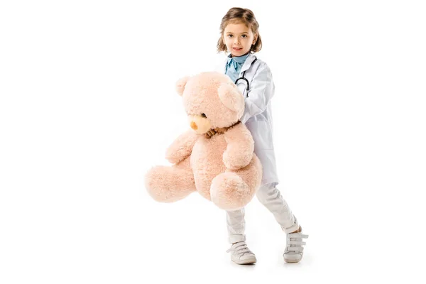 Kid dressed in doctors white coat with stethoscope holding big teddy bear isolated on white — Stock Photo