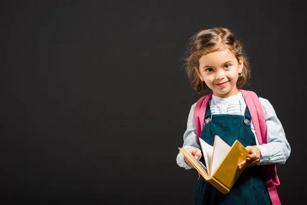 Nettes Schulmädchen mit Rucksack und Buch in den Händen, die Kamera isoliert auf schwarz — Stockfoto