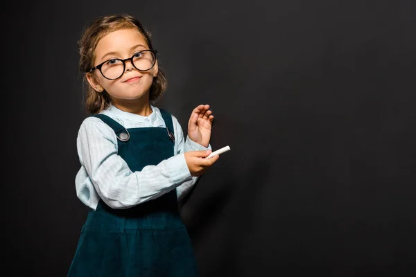 Retrato del escolar en gafas con trozo de tiza parado en pizarra en blanco - foto de stock