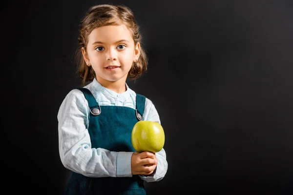 Porträt eines kleinen Schulmädchens mit Apfel in den Händen, das isoliert auf schwarz in die Kamera blickt — Stockfoto