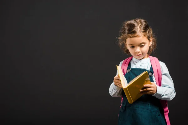 Cute schoolgirl with backpack and book in hands isolated on black — Stock Photo