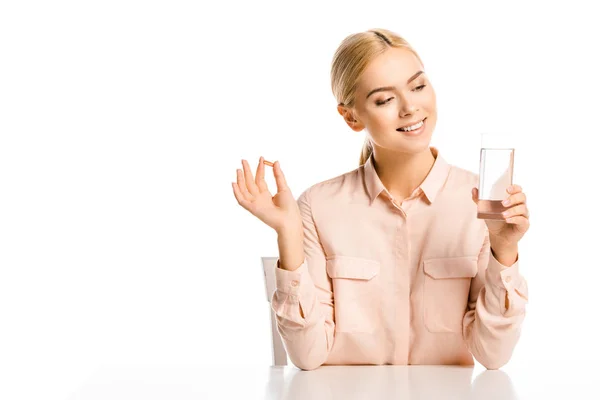 Donna attraente sorridente guardando vetro d'acqua e tenendo pillola isolata su bianco — Foto stock
