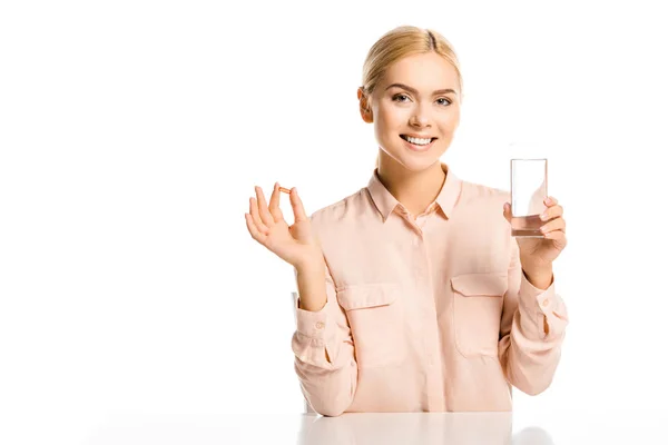 Sonriente atractiva mujer sosteniendo vaso de agua y píldora aislada en blanco, mirando a la cámara - foto de stock