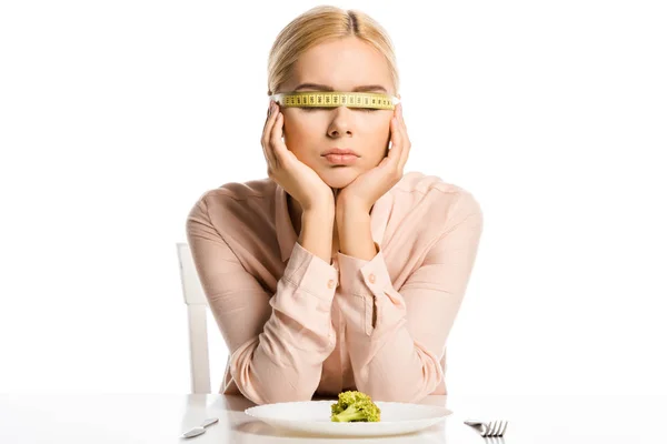Atractiva mujer con cinta métrica en los ojos sentados cerca de pedazo de brócoli en el plato aislado en blanco - foto de stock