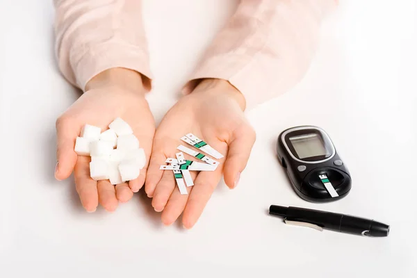 Обрезанное изображение женщины, держащей полоски глюкометра и рафинированный сахар, изолированные на белом, сахарный диабет — стоковое фото